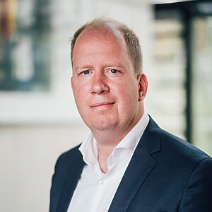 Porträt von Jan Holze, dem Vorstand der DSEE. Er blickt mit einem Lächeln direkt in die Kamera, trägt ein dunkles Jackett und ein weißes Hemd.