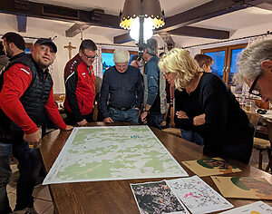 250311_Dorfwerkstatt -koimadVereint_eV_Kollnburg_Bayern_ELANetzwerk Thomas Bielmeier Menschen stehen bei einem Vereinstreffen, es gibt Bier in Bierhumpen, in einem Vereinsraum um einen Tisch auf dem mehrere kommunale Flurkarten liegen und schauen diese an.